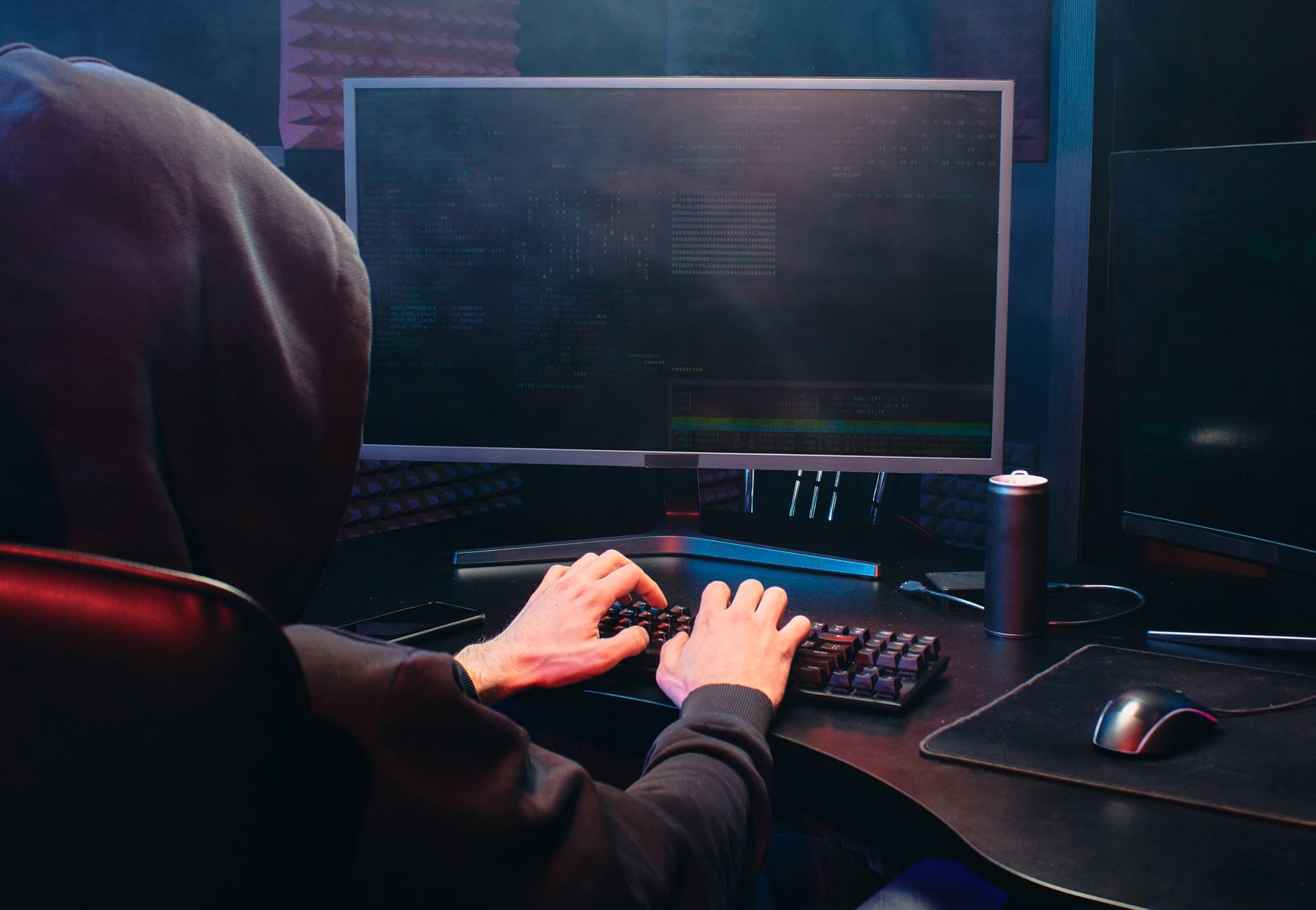 A person in a hoodie sits at a computer screen, engaged in coding or hacking activities.